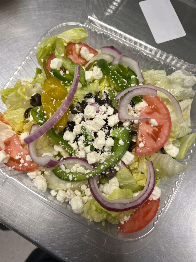 A vibrant Greek salad in a clear plastic container, featuring lettuce, tomatoes, onions, peppers, olives, and feta cheese.