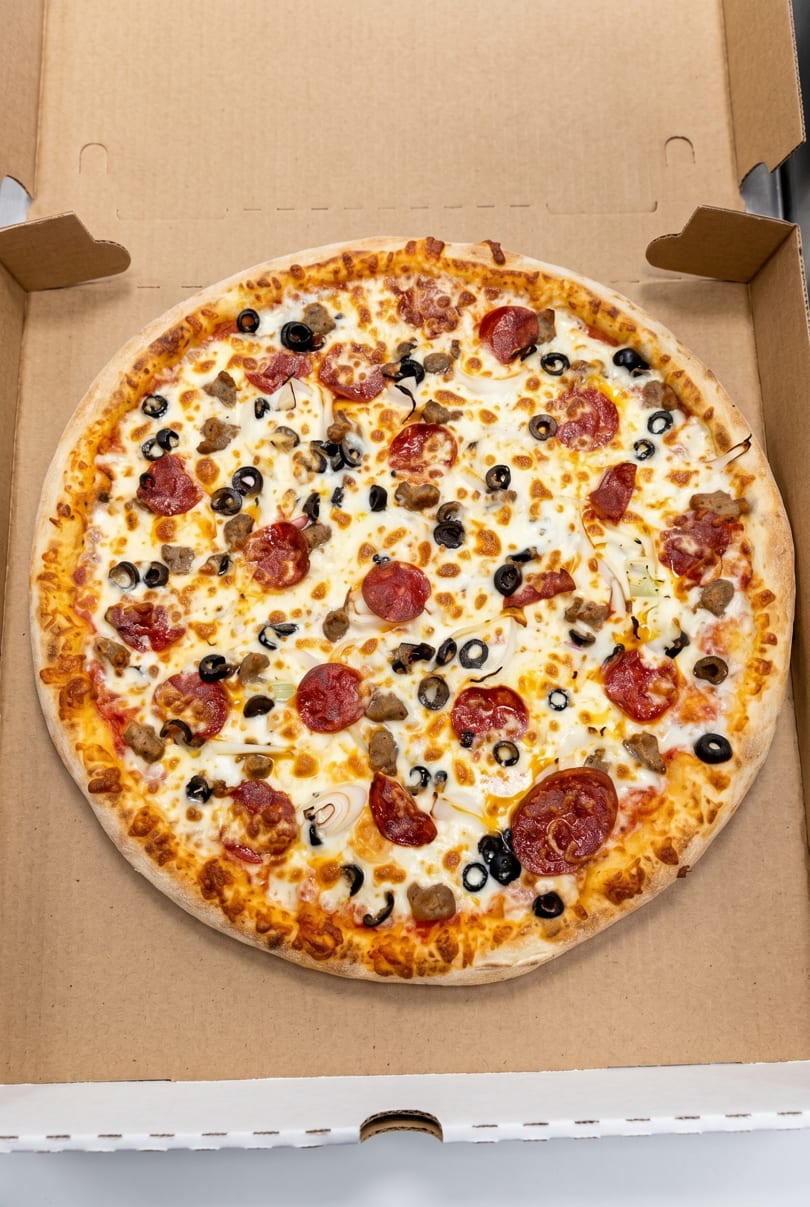 A whole pizza with pepperoni, sausage, black olives, and onions in a cardboard box.